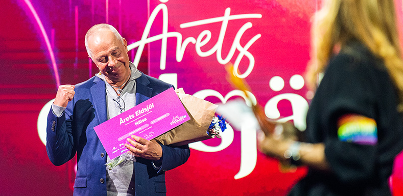 Glad man knyter ena näven, håller en check och blommor i andra handen. 